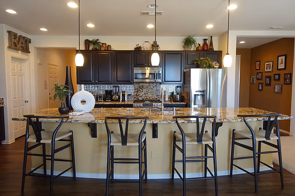 Modern kitchen with a large granite island, four bar stools, dark cabinets, stainless steel appliances and pendant lights.