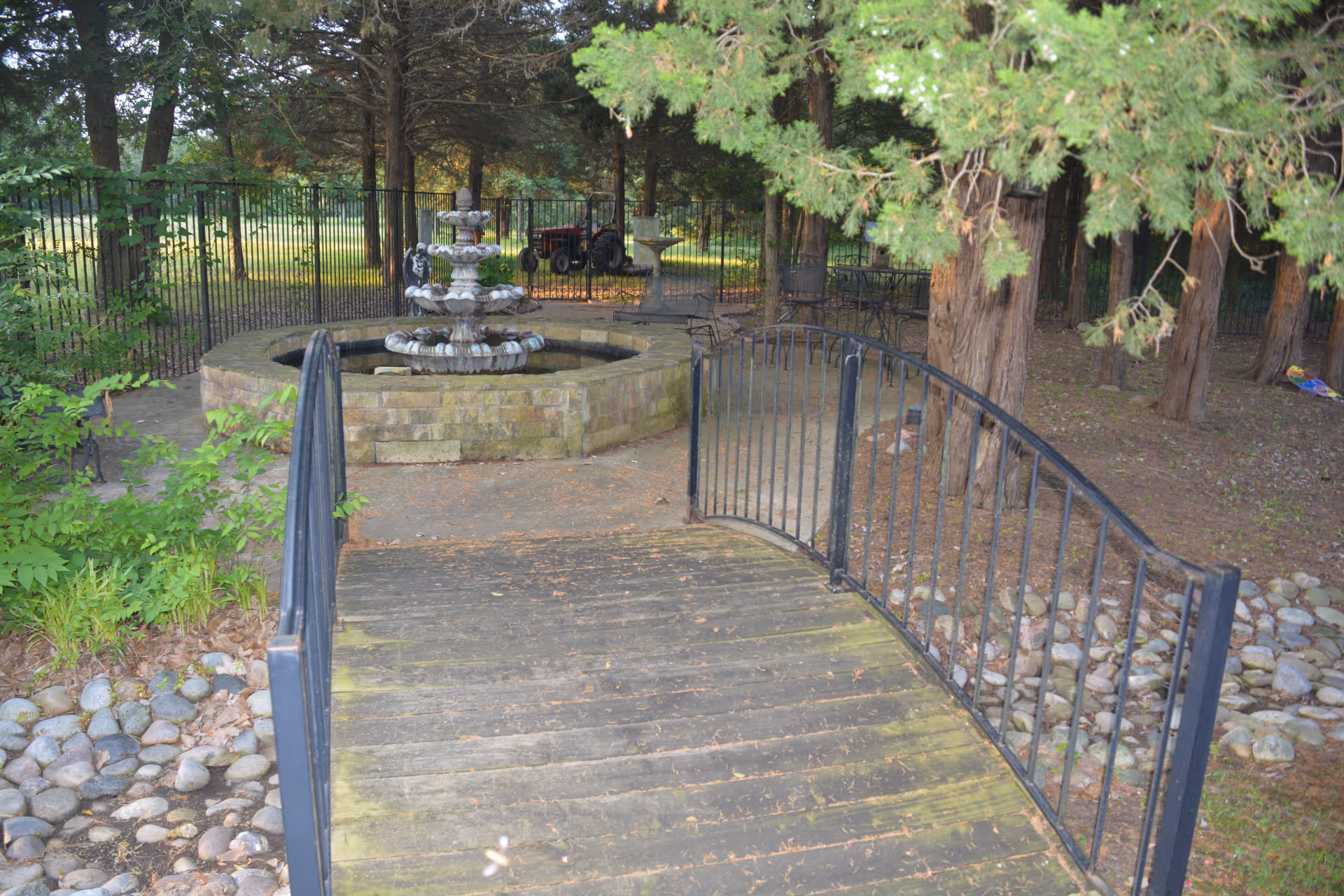 A wooden footbridge with black metal railings leads to a stone fountain in a shaded garden surrounded by trees.