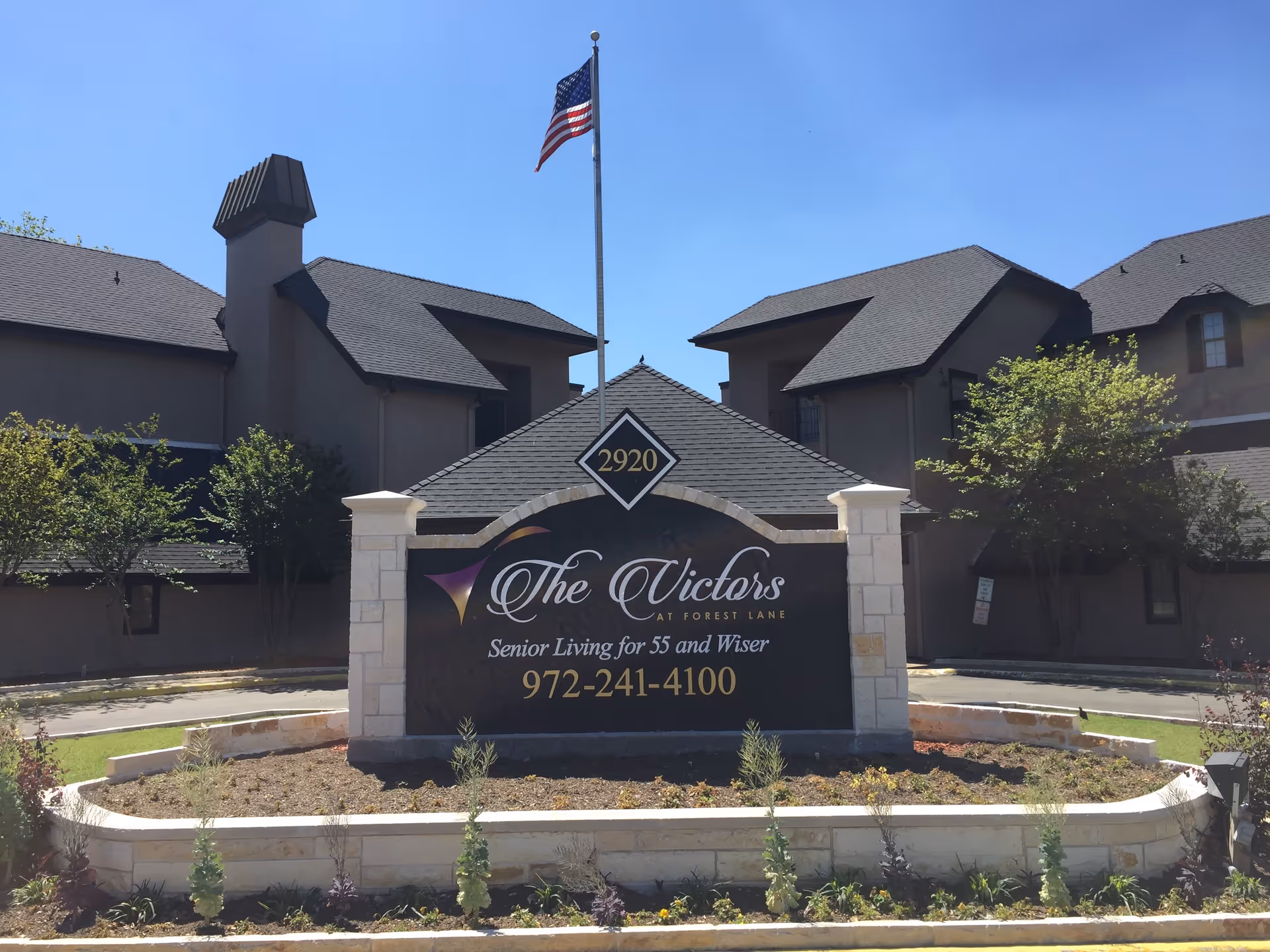 Entrance sign for The Victors senior living community in front of a multi-story building with an American flag overhead.