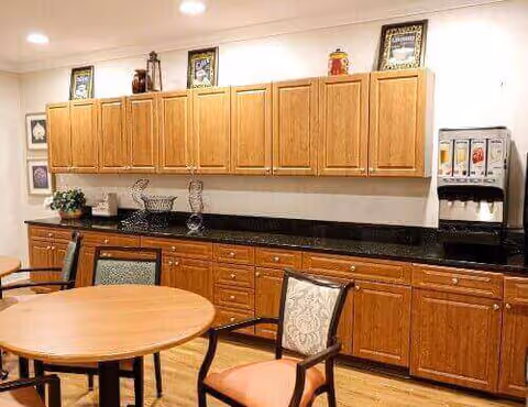 Interior view of a dining area with wooden tables and chairs. There is a long counter with wooden cabinets above and below, a black countertop, and a beverage dispenser on the right side. The walls are decorated with framed pictures and small decorative items on top of the cabinets.