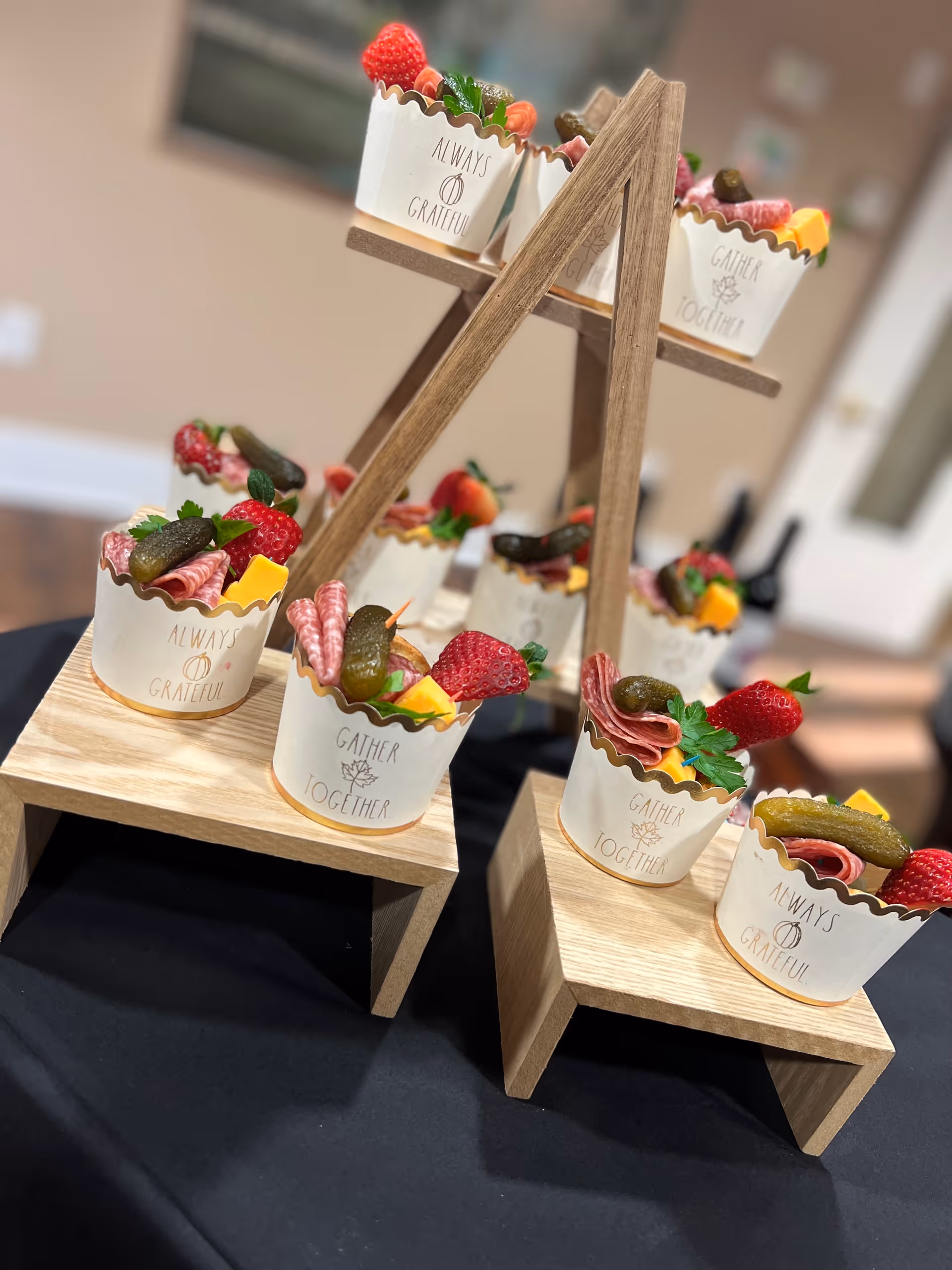 A display of small cups filled with an assortment of snacks including strawberries, pickles, rolled deli meats, cheese cubes, and herbs. The cups are white with gold scalloped edges and have phrases like 'Always Grateful' and 'Gather Together' printed on them. They are arranged on a wooden tiered stand on a black tablecloth in an indoor setting.