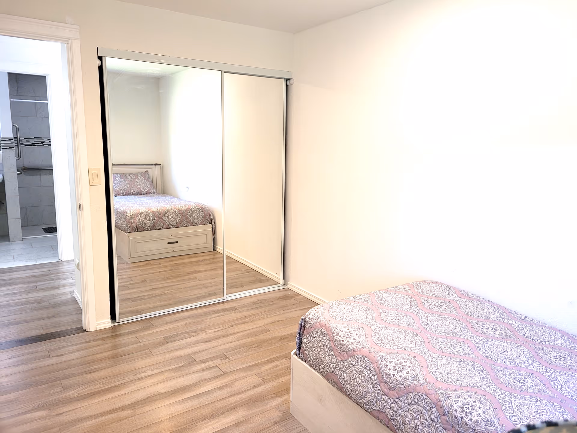 A small bedroom with light wood flooring, a bed with a patterned pink and gray bedspread, and a large closet with sliding mirrored doors. The room has white walls and a doorway leading to a bathroom with tiled walls and grab bars.