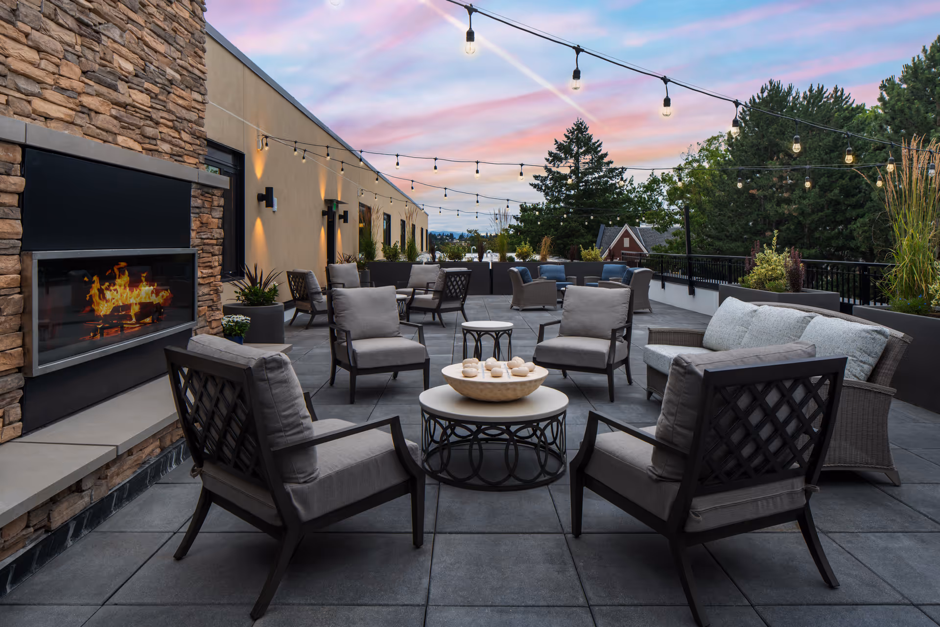 Outdoor patio area at sunset with cushioned chairs and sofas arranged around small tables. A modern stone fireplace with a fire burning is on the left side. String lights are hung overhead, and there are plants and trees in the background.