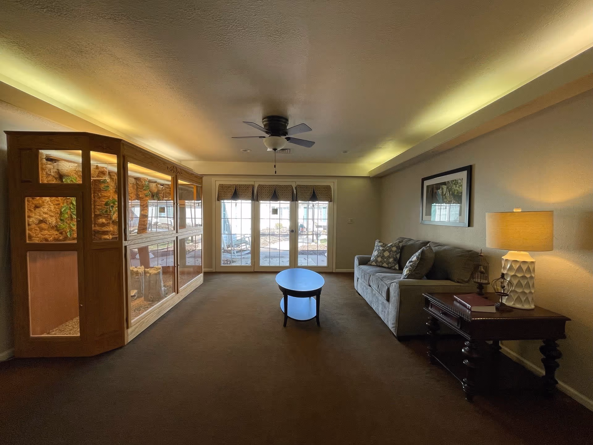 A living room with a gray couch adorned with patterned pillows, a wooden side table with a lamp and decorative items, a small oval coffee table in the center, and a large wooden display cabinet with glass panels on the left. The room has a ceiling fan and recessed lighting with a soft yellow glow. French doors with window coverings lead to an outdoor area.