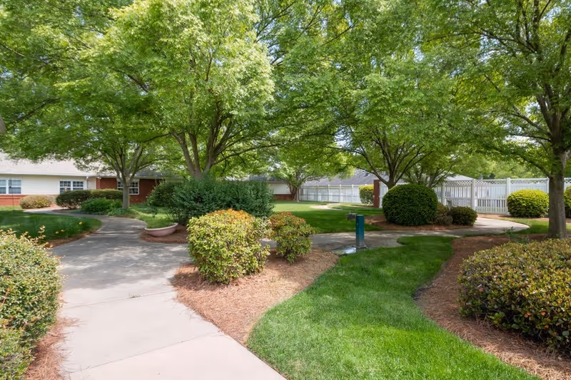 A landscaped outdoor area with a concrete walkway winding through green grass, bushes, and trees. In the background, there is a white fence and a building with white siding and red brick accents.