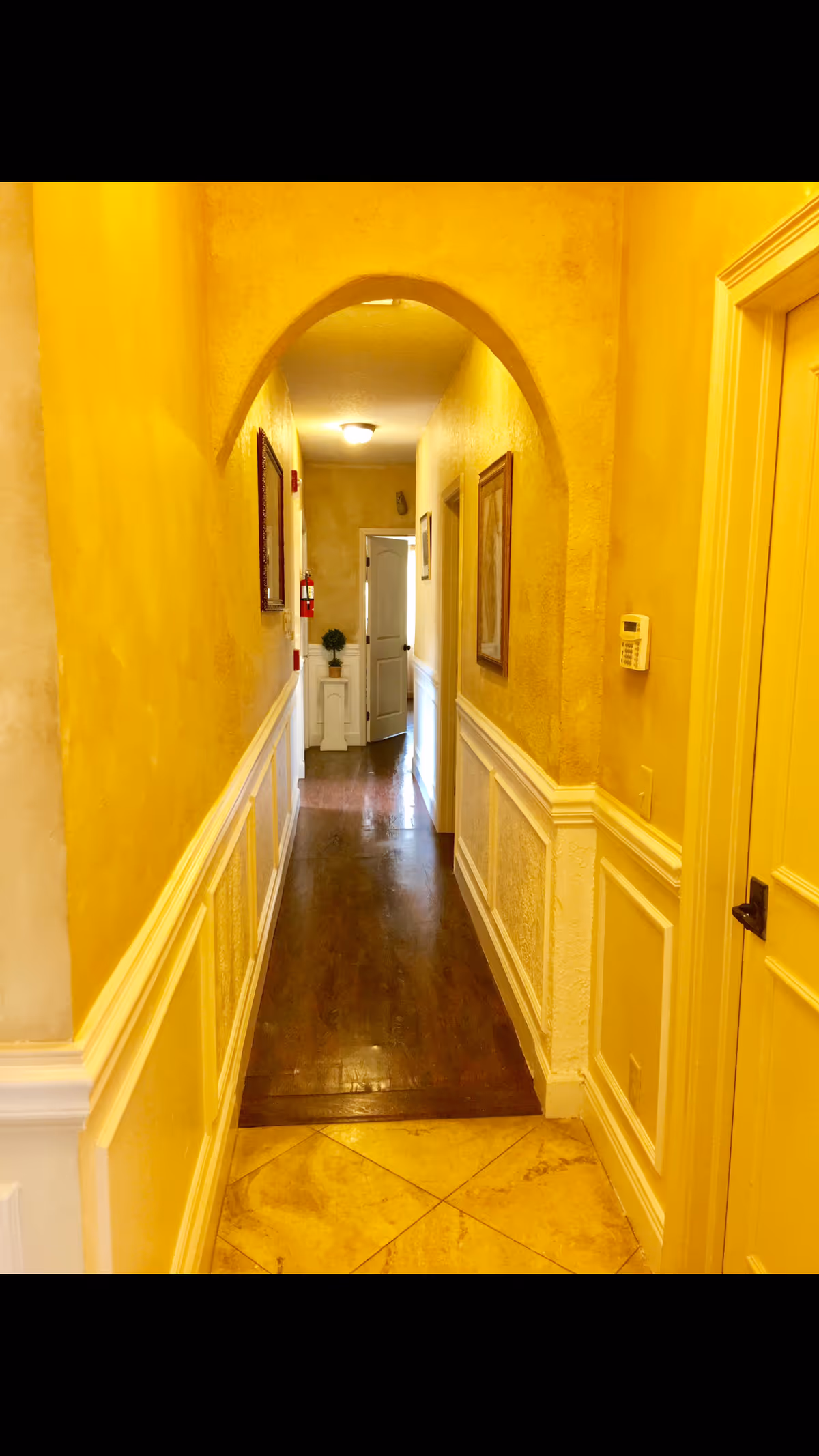 A narrow hallway with yellow textured walls and white wainscoting. The floor transitions from beige tiles to dark wood. There is an arched doorway near the middle of the hallway, framed pictures on the walls, a thermostat on the right wall, and a small white cabinet with a plant at the far end near an open door.