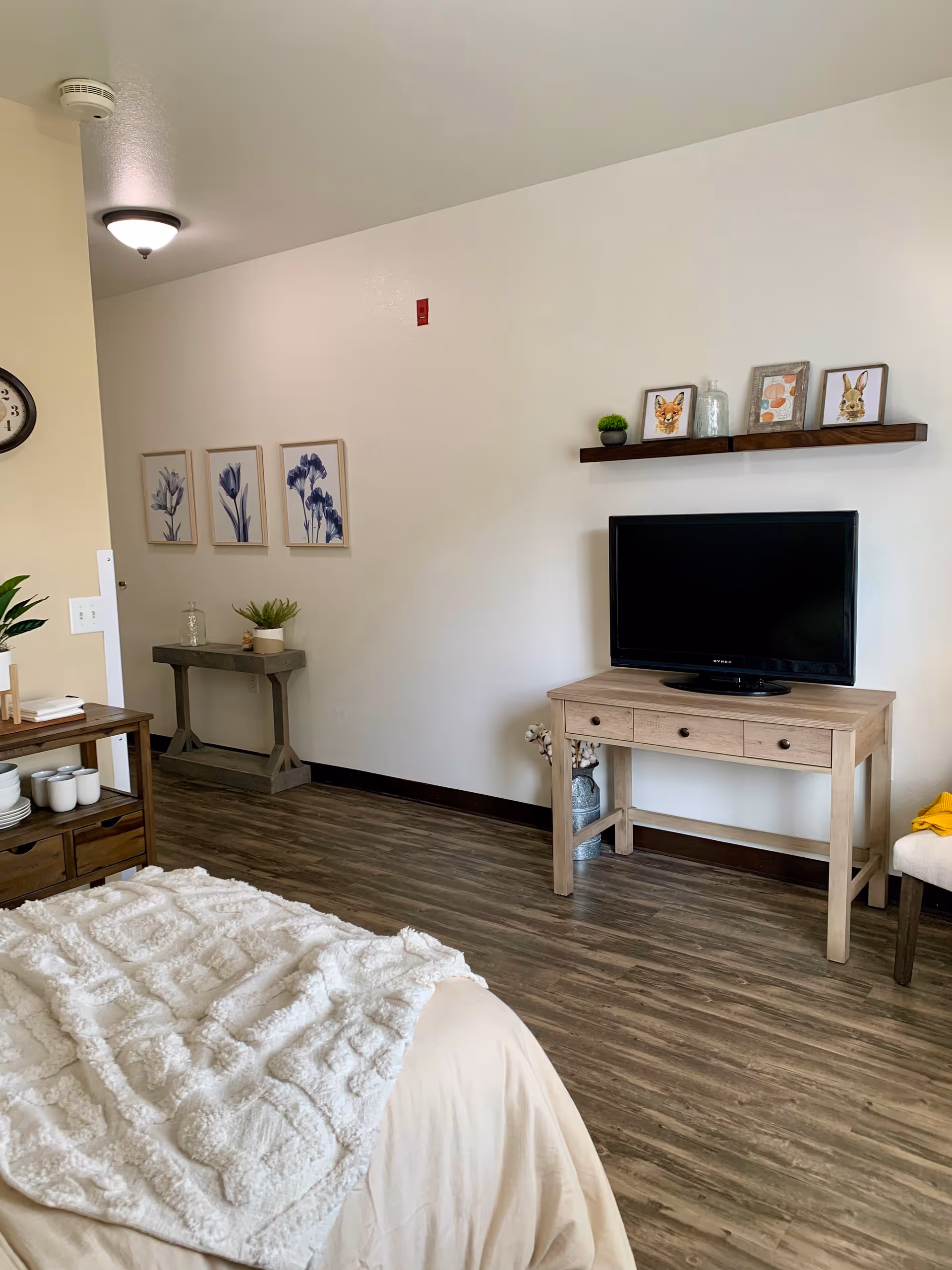 A cozy senior living bedroom with a bed covered in a beige blanket and a white textured throw. The room has wood flooring and light-colored walls. A wooden console table holds a flat-screen TV, with a decorative shelf above displaying framed pictures and a small plant. On the left side, there is a small table with a plant and glass jar, and three framed floral artworks hang on the wall. A clock is partially visible on the left wall.