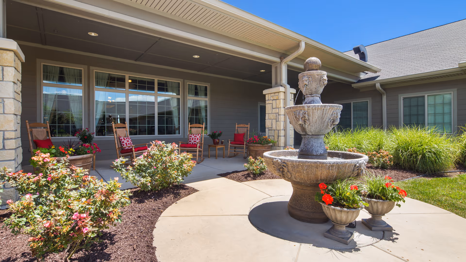 Outdoor patio area at Regent Park Assisted Living & Memory Care featuring a three-tier stone fountain surrounded by flower pots with red flowers. The patio has several wooden rocking chairs with red cushions and small tables, set against a building with large windows and stone pillars. The area is landscaped with bushes and grass under a clear blue sky.