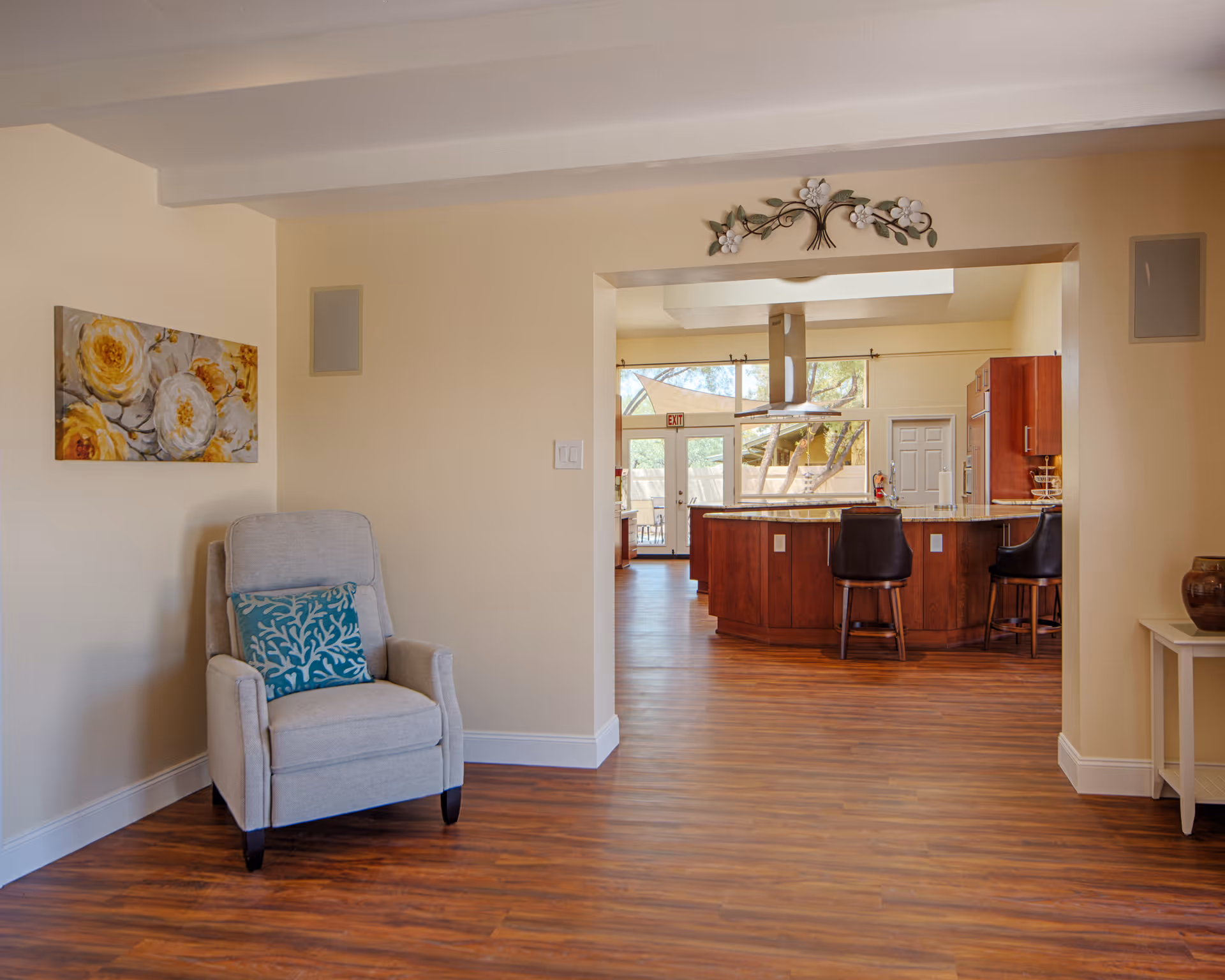 Interior view of a senior living facility showing a cozy seating area with a gray armchair and a decorative pillow, a floral painting on the wall, and a kitchen area in the background with wooden cabinetry, a kitchen island with two black bar stools, and large windows letting in natural light.