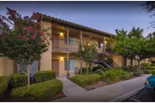Exterior view of a two-story apartment building at dusk with a walkway, trees, and shrubs in front. The building has a balcony on the second floor and exterior stairs leading up. Warm lights illuminate the entrances and balcony area.