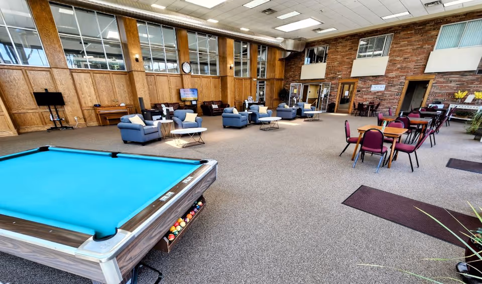 Spacious communal lounge with a pool table, multiple seating areas, and dining tables in a senior living facility.