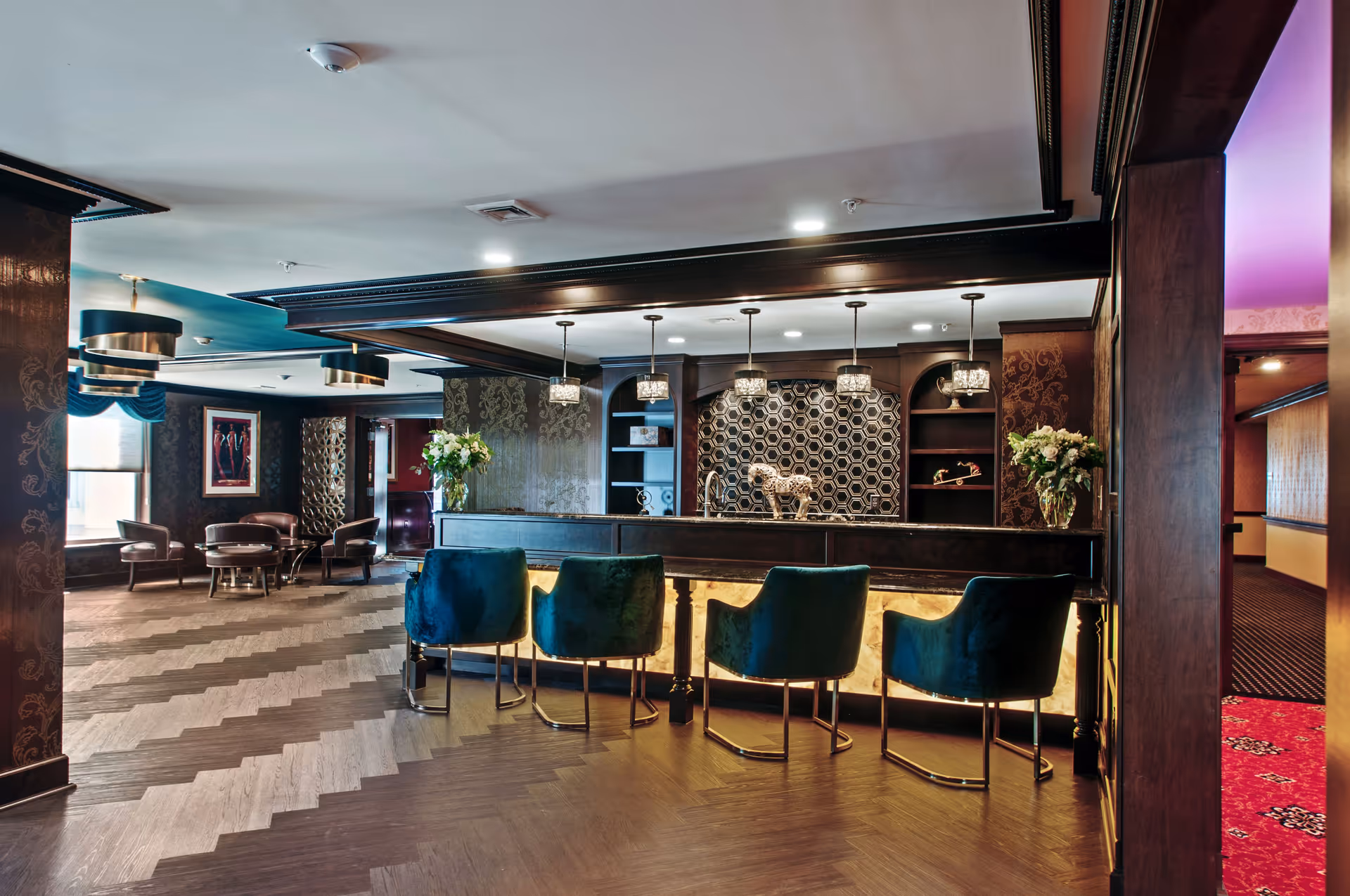 Interior view of a stylish lounge area with a bar counter featuring four teal velvet chairs. The bar has a decorative honeycomb patterned backsplash and pendant lights hanging above. To the left, there is a seating area with round tables and chairs near windows with blue valances. The space has dark wood accents, patterned wallpaper, and a chevron-patterned floor.