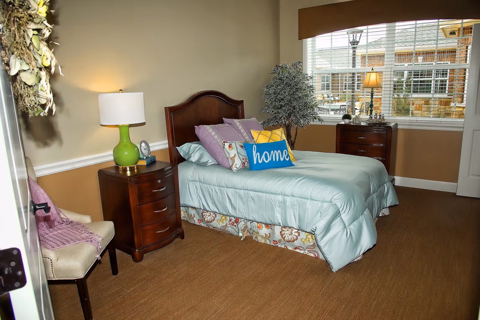 A cozy bedroom with a single bed dressed in light blue bedding and multiple decorative pillows, including one with the word 'home'. The room features a wooden nightstand with a green lamp and a clock, a beige chair with a purple throw, a wooden dresser near a large window with blinds, and a potted plant. The walls are painted beige with white trim, and the floor is carpeted in brown.