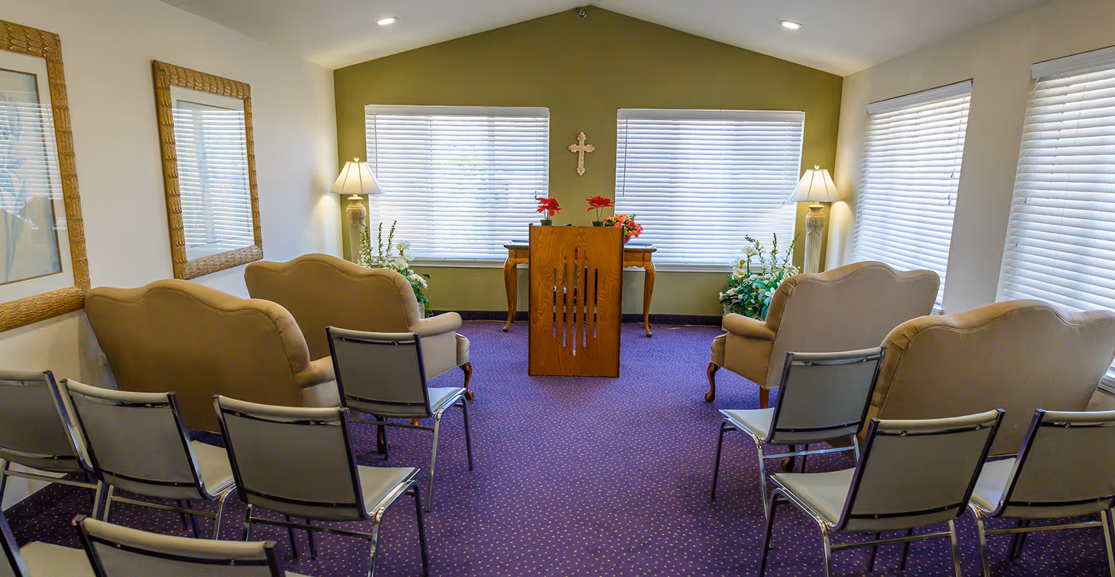 Small chapel-style meeting room with a wooden lectern, upholstered armchairs and rows of metal chairs facing windows.