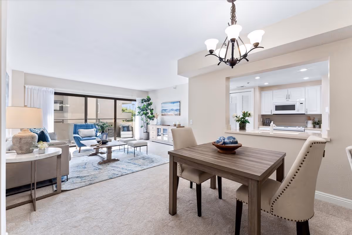 A bright and spacious living and dining area in a senior living facility. The living room features a beige sofa, a blue armchair, a wooden coffee table, and a large window with sliding glass doors leading to a balcony. The dining area has a wooden table with two upholstered chairs and a decorative bowl. The kitchen is visible through a pass-through window and includes white cabinets, a microwave, and a stove. The room is decorated with plants, artwork, and a chandelier above the dining table.