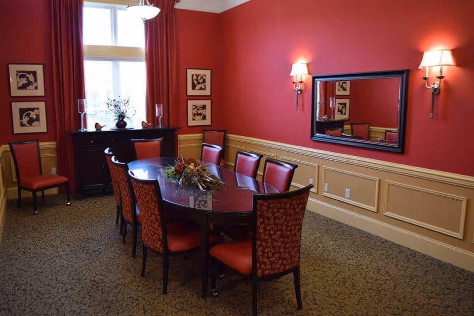 A formal dining room with a long oval wooden table surrounded by eight red cushioned chairs. The walls are painted red with beige wainscoting, decorated with framed artwork and a large rectangular mirror. Two wall sconces with lampshades provide lighting, and a window with red curtains lets in natural light. A dark wooden sideboard with decorative items is positioned under the window.