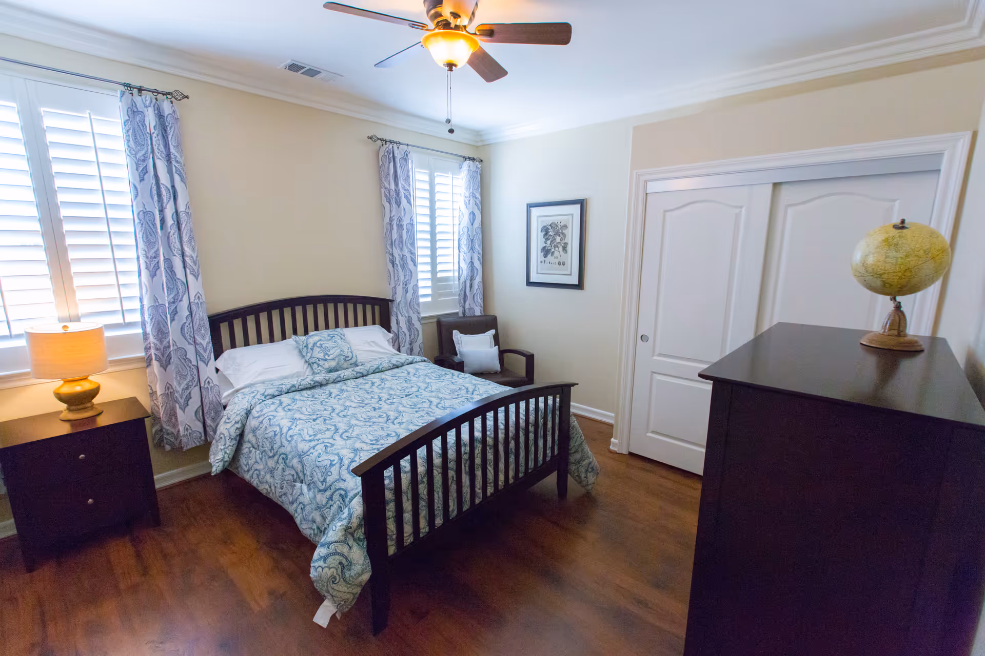 A cozy bedroom with a wooden bed frame and a blue patterned bedspread. There are two windows with white shutters and blue patterned curtains. A nightstand with a lamp is beside the bed, and a wooden dresser with a globe on top is against the opposite wall. A framed picture hangs on the wall next to a sliding closet door.