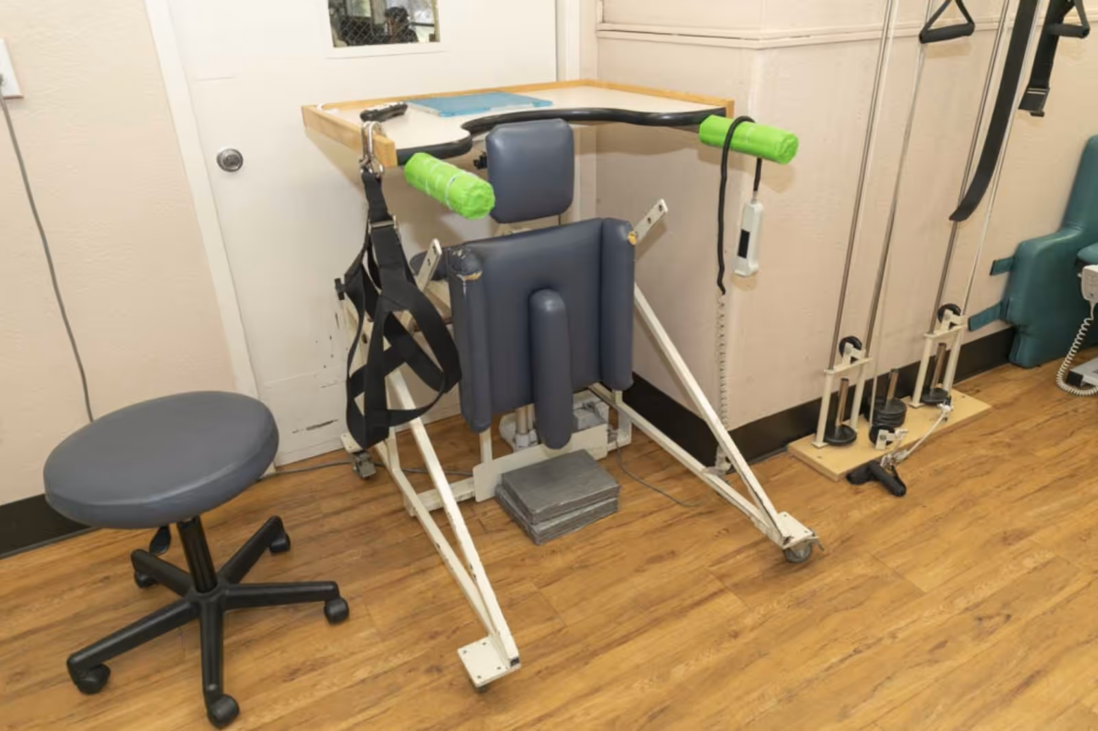 A physical therapy or rehabilitation room featuring exercise equipment including a padded chair with green handles, straps, pulleys, and a rolling stool on a wooden floor.