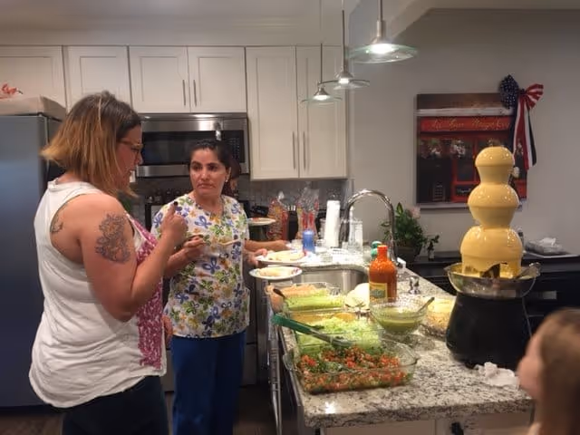 Two women standing in a kitchen with a granite countertop island. One woman is holding a plate and appears to be serving herself food from several dishes on the counter, including a large yellow chocolate fountain. The kitchen has white cabinets, a stainless steel refrigerator, and a microwave. A child is partially visible in the foreground.