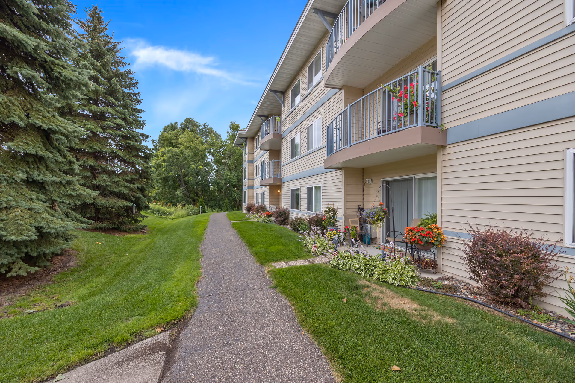 Exterior view of a multi-story residential building with beige siding and balconies. There is a paved walkway alongside the building, bordered by well-maintained green grass and various shrubs and flowers. Tall evergreen trees and other greenery are visible in the background under a partly cloudy blue sky.