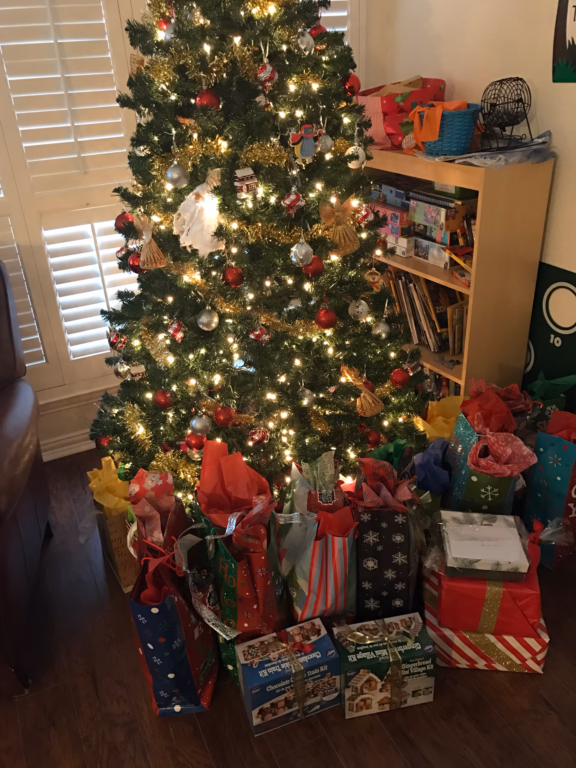 A decorated Christmas tree with white lights, red and silver ornaments, and gold tinsel in a room with wooden flooring. Numerous wrapped gifts and gift bags are placed under the tree. A wooden bookshelf filled with books and games is visible in the background, along with a bingo cage on top of the shelf.