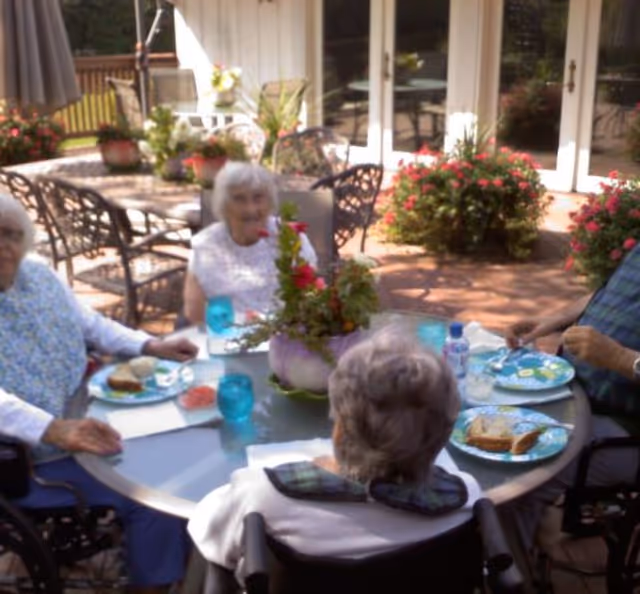 Four elderly people sitting around a round outdoor table having a meal together. The table has blue glasses, plates with sandwiches, and a floral centerpiece. The setting is a patio with potted plants and glass doors leading inside.