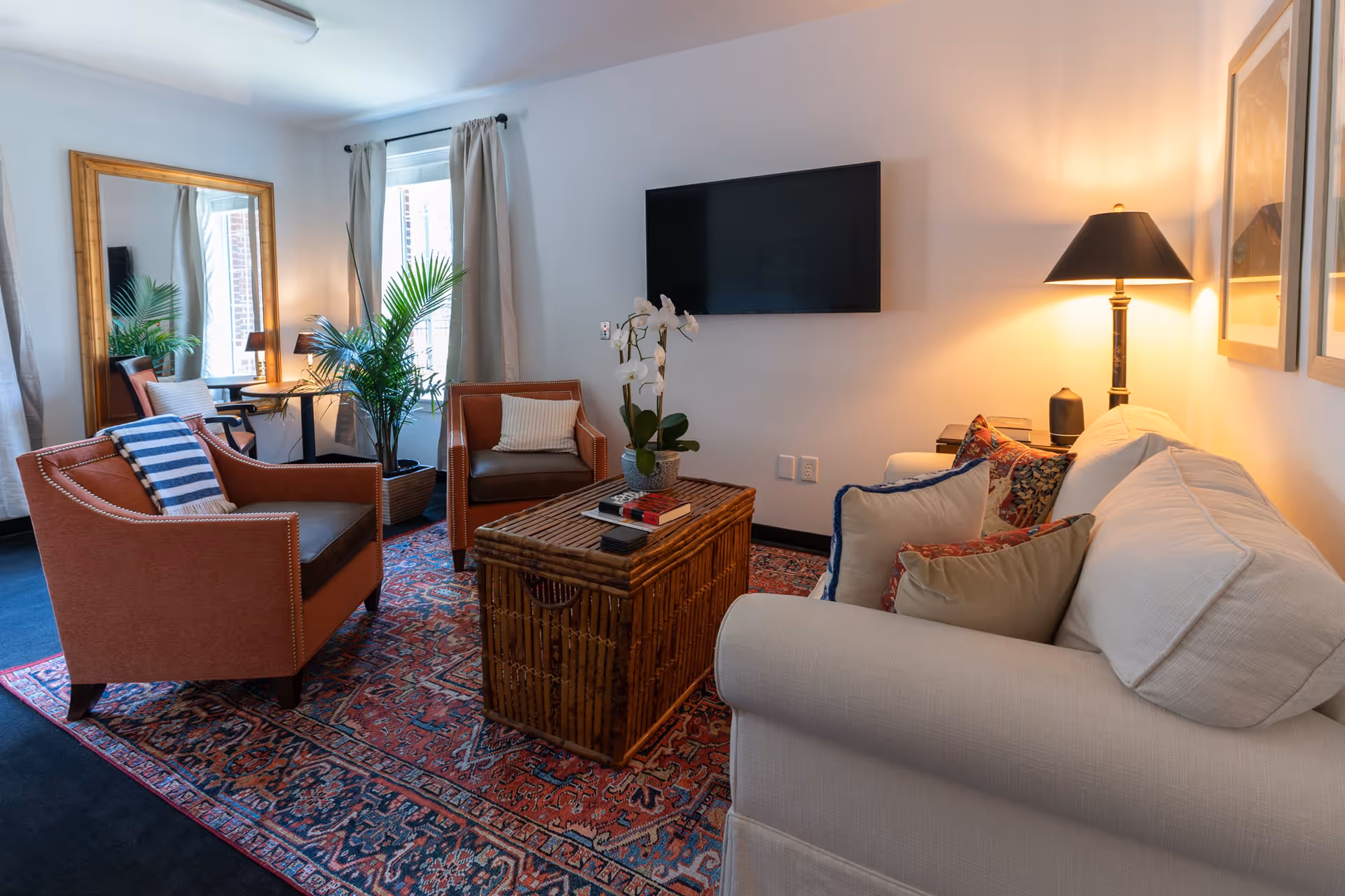 A cozy living room with a white sofa adorned with multiple pillows, two brown armchairs with cushions, a wooden coffee table with books and a potted orchid, a large mirror leaning against the wall, a potted plant near the window with curtains, a floor lamp, and a flat-screen TV mounted on the wall.