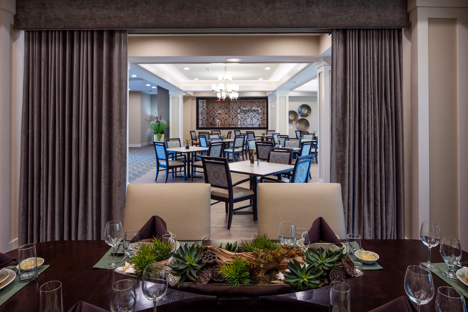View of a dining area in a senior living facility with a dark wooden table set with glassware, plates, and napkins in the foreground. The table has a centerpiece with green succulents and decorative elements. Beyond the table, there is a larger dining room with multiple tables and chairs, soft lighting, and decorative wall art.