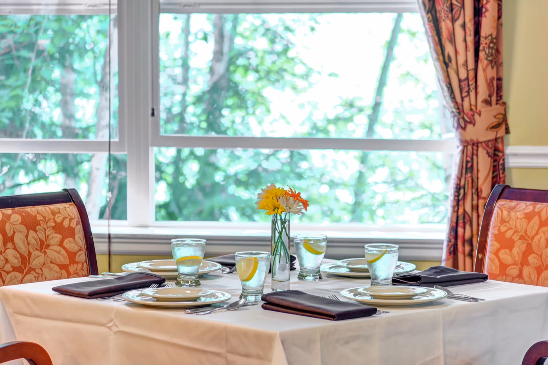 A dining table set for four with white tablecloth, plates, glasses of water with lemon slices, black napkins, and a small vase with yellow and orange flowers. The table is positioned in front of a large window with a view of green trees outside. The chairs have orange floral upholstery and there are patterned curtains on the side.