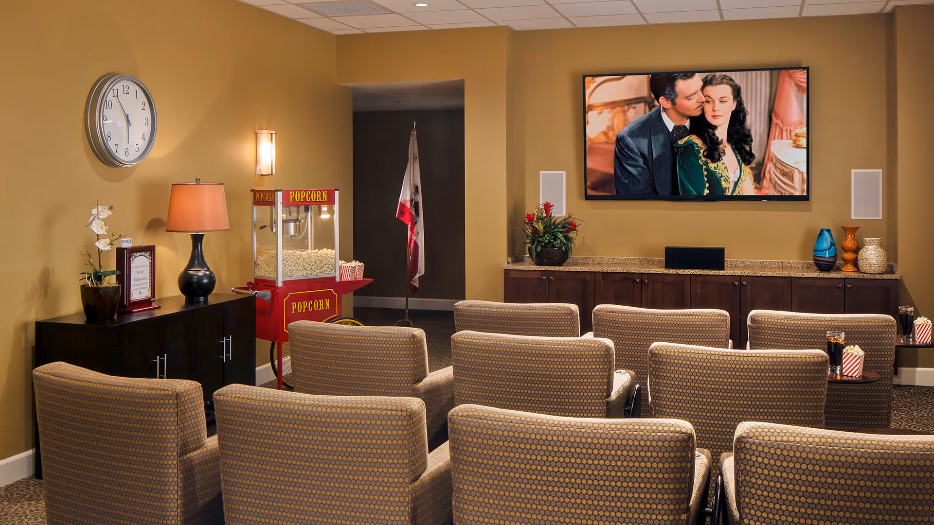 A cozy movie theater room with several rows of cushioned chairs facing a large flat-screen TV mounted on a beige wall. A popcorn machine filled with popcorn is positioned near a black cabinet with a lamp and decorative items. The room has warm lighting and a carpeted floor.
