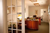 View through glass-paneled double doors into an office area with a wooden desk, computer, chair, and decorative plants. The room has beige walls, framed artwork, and ceiling light fixtures. A white door is visible at the end of the hallway.