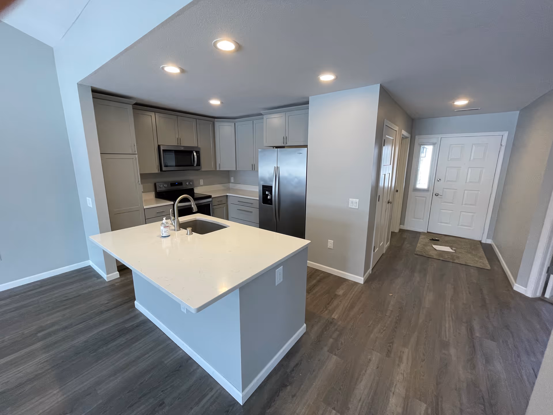 Modern kitchen with gray cabinets, stainless steel refrigerator, microwave, and stove. A white island with a built-in sink and faucet is in the foreground, with a bottle of hand soap on the countertop. The kitchen opens to a hallway with a white front door and wood flooring throughout.
