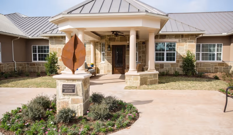 Front entrance of The Ridge at Westover facility featuring a stone building with a covered porch supported by columns, a wooden door, windows with white frames, and a landscaped area with a metal leaf sculpture on a stone pedestal.