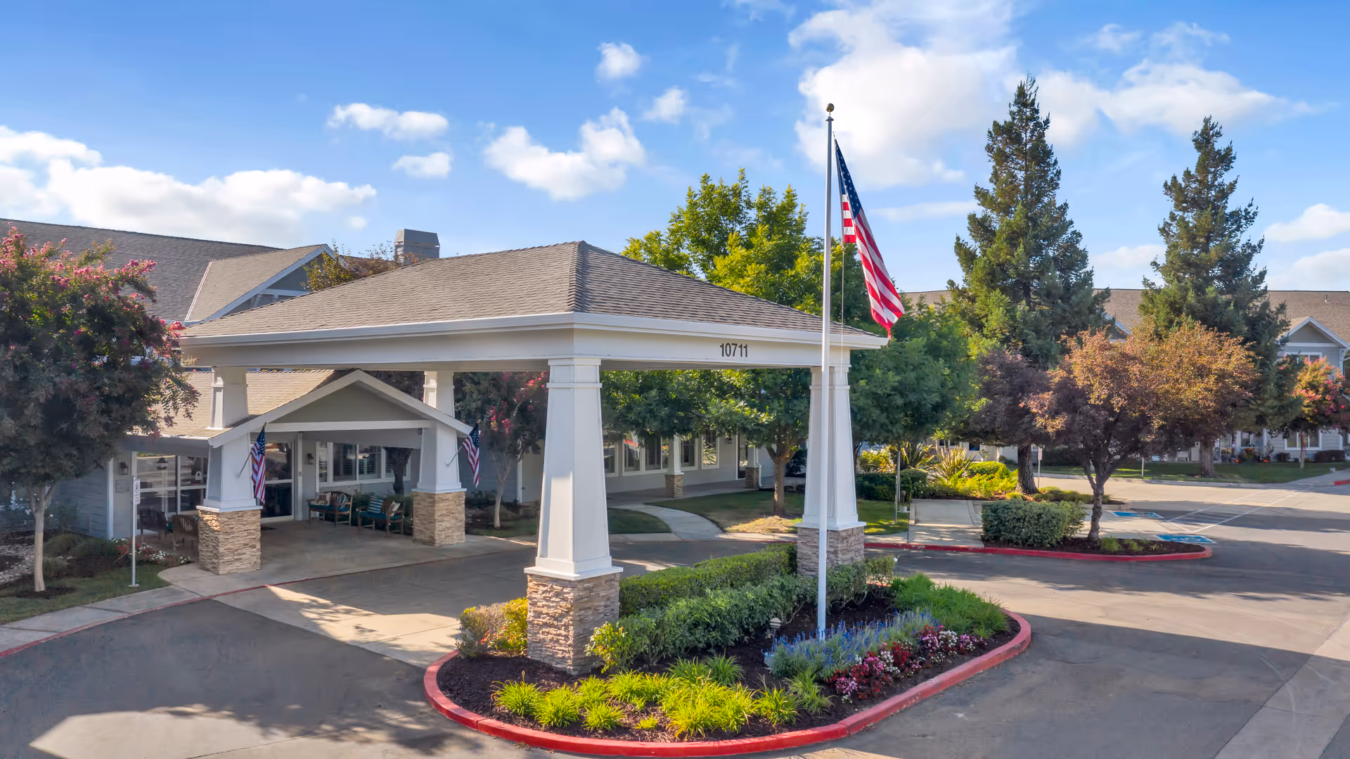 Exterior view of The Commons on Thornton senior living facility entrance with a covered driveway, American flag on a flagpole, landscaped greenery, and trees under a partly cloudy sky.
