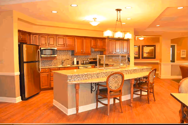 A warm and inviting kitchen area with wooden cabinets, stainless steel refrigerator, microwave, and stove. There is a tiled countertop island with two wooden chairs featuring patterned cushions. The room has hardwood flooring and soft yellow lighting from ceiling fixtures and a hanging light above the island. The kitchen opens into a hallway with framed artwork on the walls.