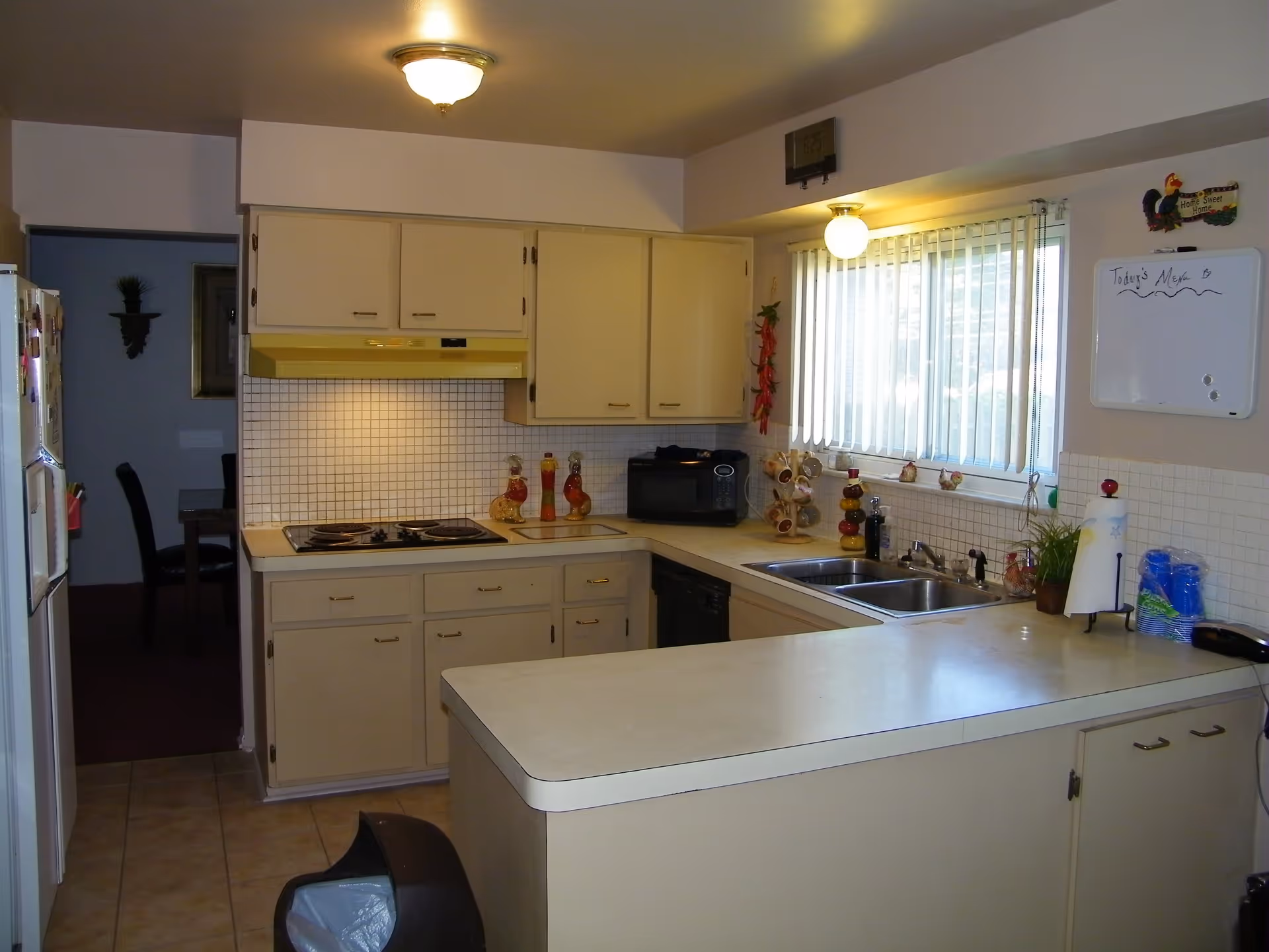 Well-lit kitchen with beige cabinets, a stove and microwave, double sink, and a countertop peninsula beneath a window with vertical blinds.