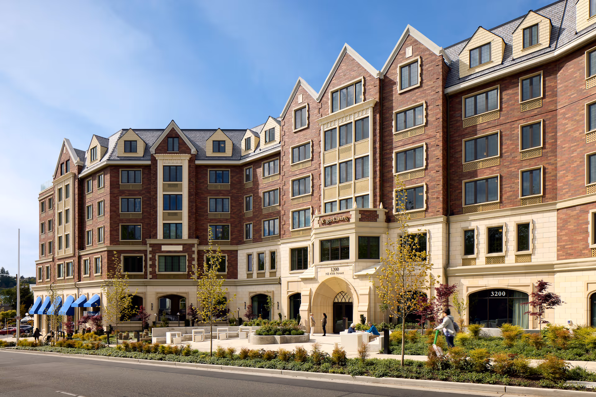 Exterior view of Aegis Living Laurelhurst, a multi-story senior living facility with brick and cream-colored facade, large windows, and a landscaped entrance area with benches and small trees. Several people are visible near the entrance and on the sidewalk.