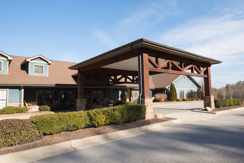 Front entrance of a single-story building with a large covered porte-cochere, landscaped shrubs, and a clear blue sky.