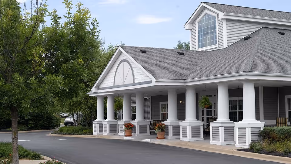 Exterior view of a senior living facility building with gray siding, white columns, and a covered entrance. There are potted plants with flowers near the entrance and trees along the driveway.