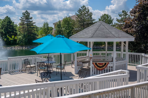A lakeside outdoor wooden deck with a white gazebo, turquoise umbrellas, and patio tables and chairs overlooking a pond and fountain.