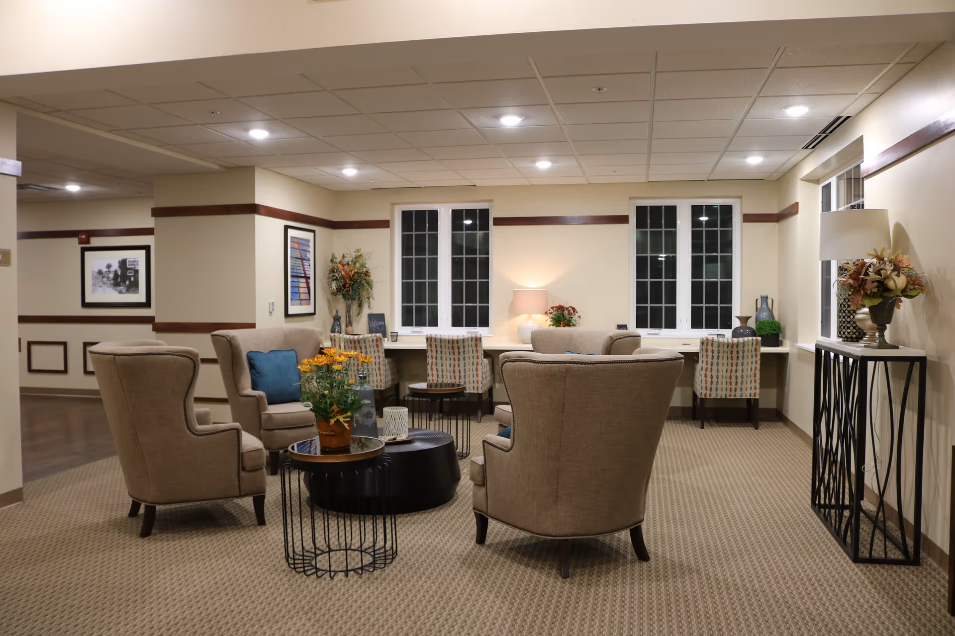 A cozy seating area in a supportive living facility with four beige upholstered armchairs arranged around a black coffee table and a small side table with a potted plant. Behind the seating area, there is a long desk with three patterned chairs, decorative vases, a lamp, and floral arrangements. The room has beige walls with wooden trim and carpeted flooring, illuminated by recessed ceiling lights.