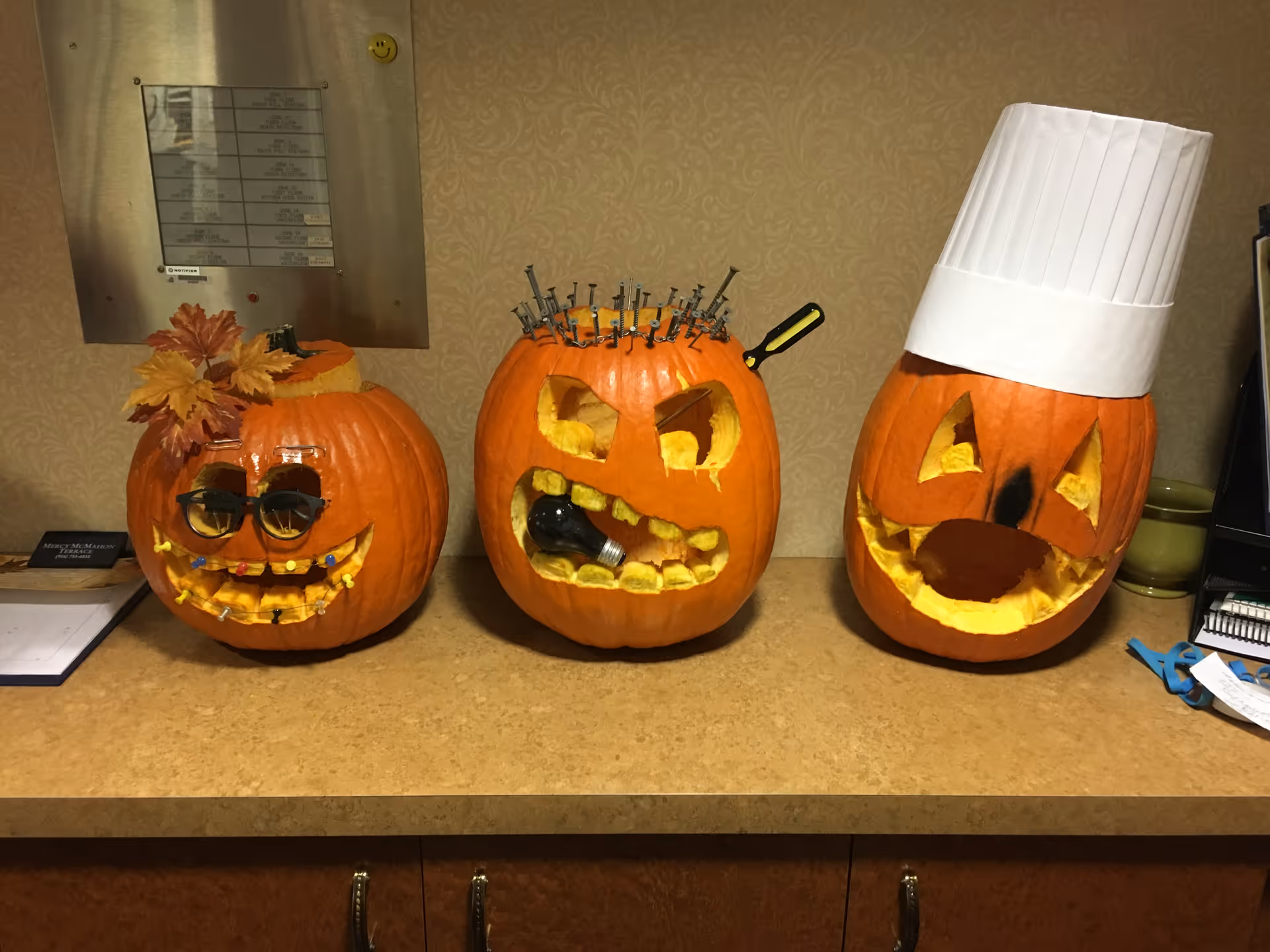 Three creatively carved pumpkins displayed on a countertop. The left pumpkin has glasses, a leafy decoration on top, and a smiling face with teeth made from small objects. The middle pumpkin has an angry expression with nails and screws stuck into the top and a light bulb in its mouth. The right pumpkin wears a white chef's hat and has a smiling face with a large nose.