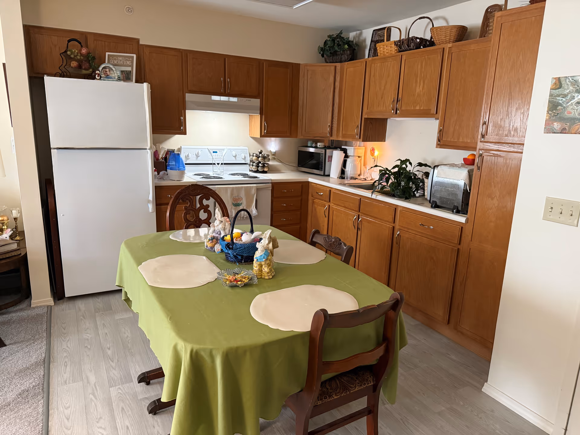 A kitchen area with wooden cabinets, a white refrigerator, stove, microwave, coffee maker, and toaster. In the foreground, there is a dining table covered with a green tablecloth and four beige placemats. The table is decorated with small bunny figurines and a basket with colorful eggs. The floor has light wood-style flooring, and there is a small piece of artwork on the wall to the right.