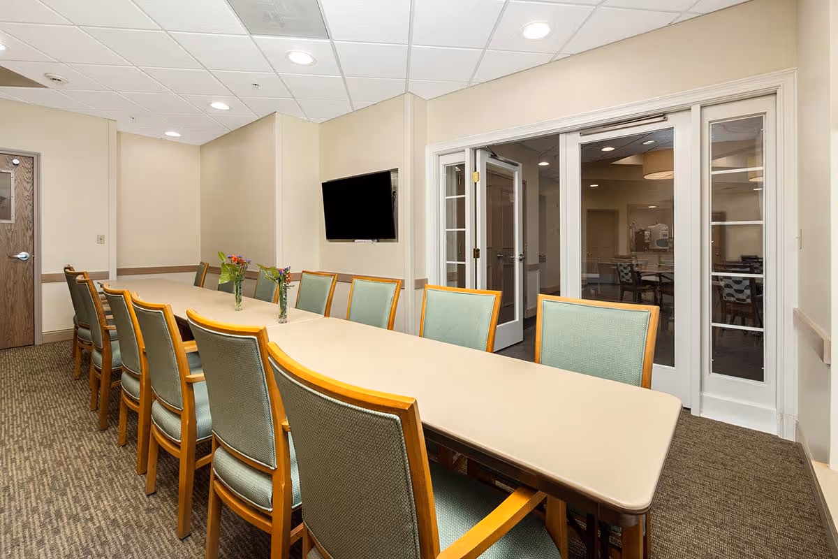 A conference or meeting room with a long beige table surrounded by green cushioned wooden chairs. There are three small vases with flowers on the table. A flat-screen TV is mounted on the wall, and there are glass double doors leading to another room. The room has beige walls, carpeted floor, and a drop ceiling with recessed lighting.