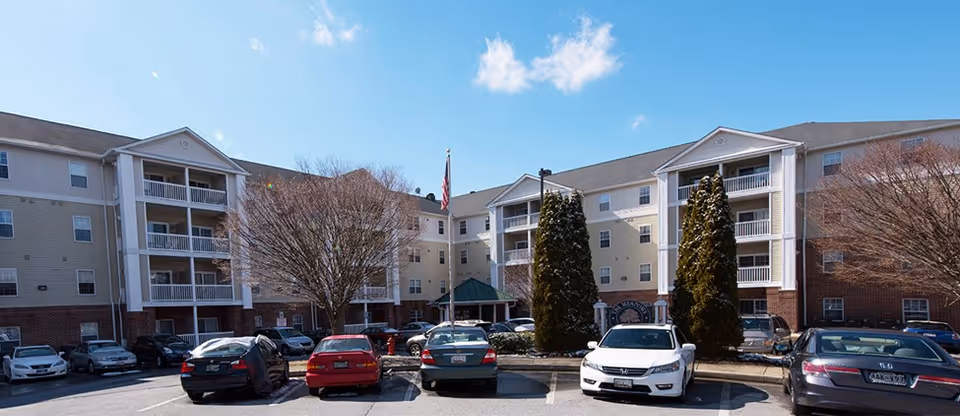 Exterior view of The Meadows of Reisterstown I & II senior living facility showing a multi-story building with balconies, several parked cars in the parking lot, leafless trees, and a clear blue sky with a few clouds.