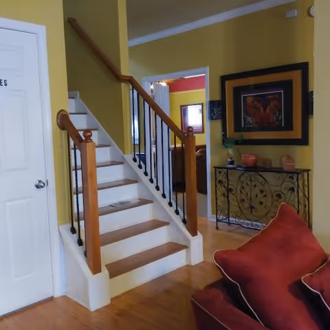 Interior view of a living area with a staircase featuring wooden handrails and black balusters. The walls are painted yellow, and there is a decorative console table with a plant and artwork above it. A red cushioned chair is partially visible in the foreground.