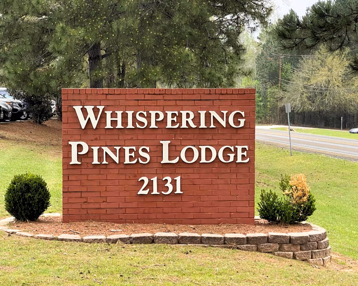 A red brick sign with white letters that reads 'Whispering Pines Lodge 2131' situated on a grassy area with small bushes and trees in the background near a road.