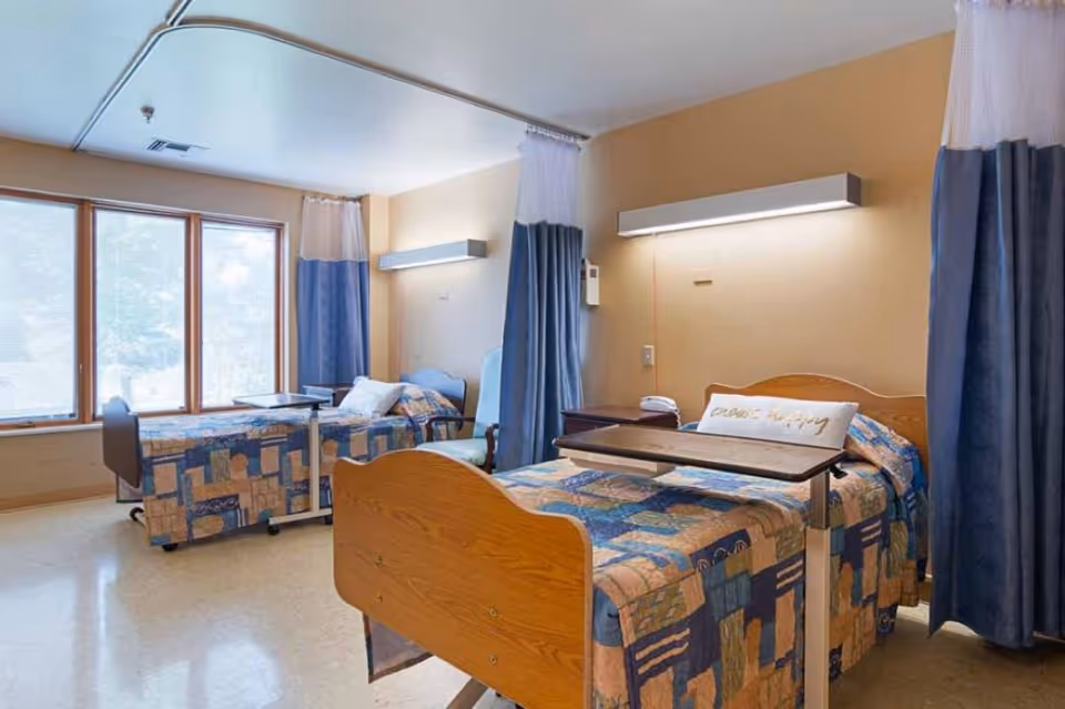 A healthcare facility room with two hospital beds separated by blue and white privacy curtains. Each bed has a patterned quilt and an overbed table. There is a large window letting in natural light and a chair next to one of the beds. The walls are beige with mounted lights above each bed.