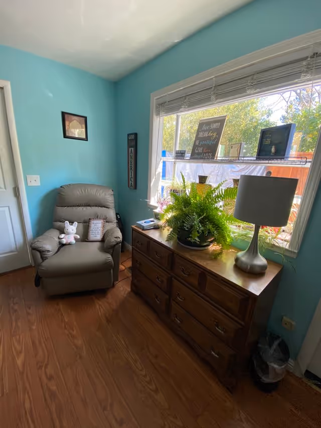 A cozy room with light blue walls featuring a comfortable beige recliner chair with a stuffed bunny and a small pillow on it. Next to the chair is a wooden dresser with multiple drawers, topped with a green potted fern, a silver lamp, and several framed inspirational quotes placed on the windowsill. The window shows a view of trees and outdoor greenery. The floor is wooden, and there is a small trash can near the dresser.