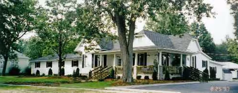 White, single-story building with a wraparound porch, trees, and a driveway.